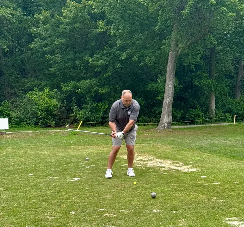 Man in light gray sports gear swinging to hit a golf ball.